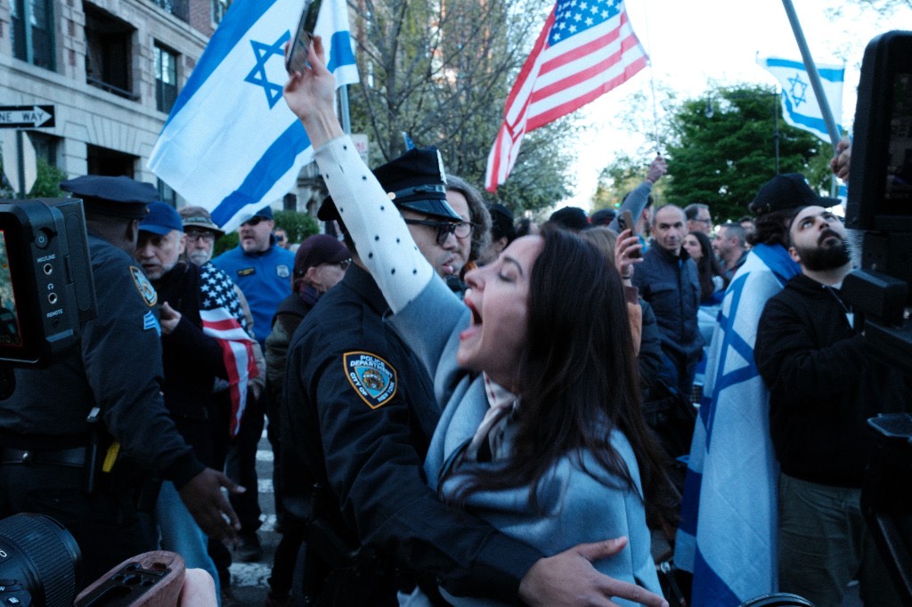 A woman shouts while holding up her phone, surrounded by Israeli flags and an NYPD officer