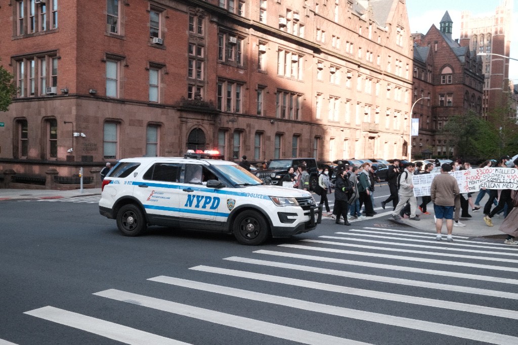 An NYPD SUV with lights on as marchers pass through a Morningside Heights intersection
